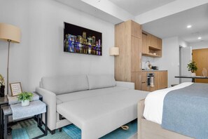 Ein modernes Hotelzimmer mit Sofa, Bett, Schreibtisch mit Lampe, einem Stadtbild als Gemälde und einer Küchenzeile.