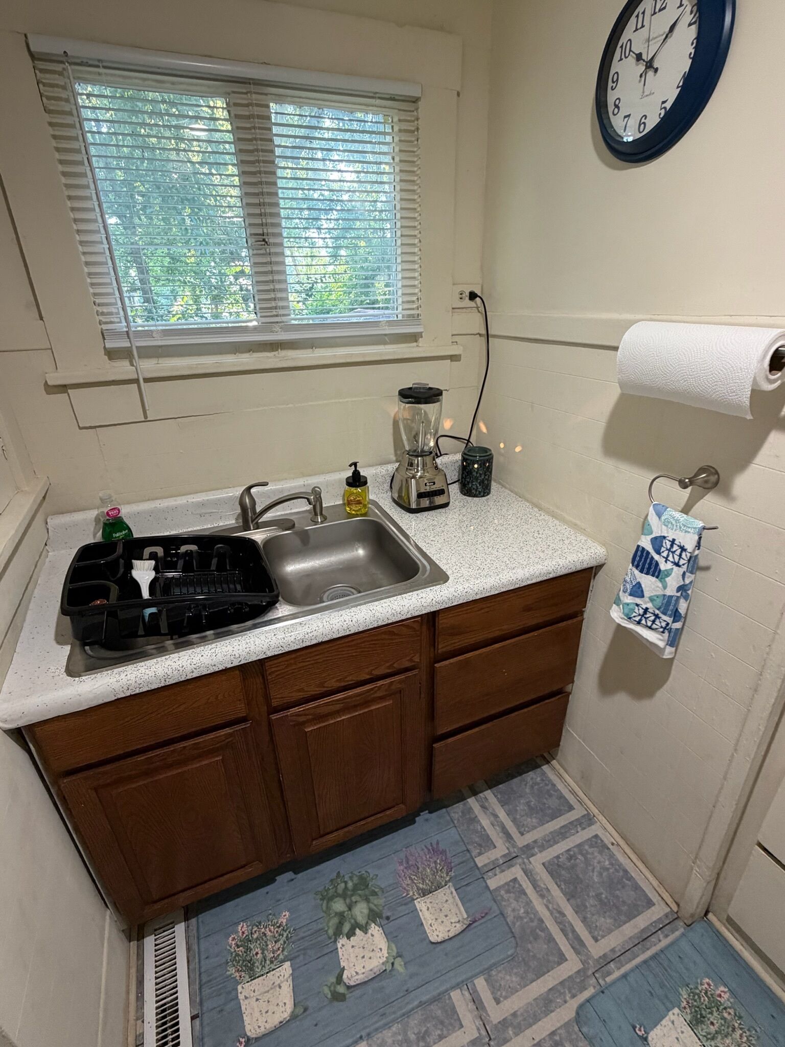A small kitchen with a sink, a blender, a window with blinds, a clock, and a towel rack.