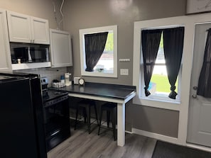 A compact kitchen with a black refrigerator, a microwave, and a coffee maker on the counter. There are two bar stools and a window with a curtain.