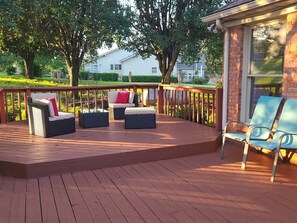 Una terraza de madera con mobiliario exterior, incluyendo sofás y sillones, y un ventanal de vidrio que da a una zona cubierta.