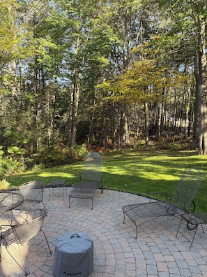 A patio with metal chairs and a round table, surrounded by a wooded area. - Beautiful home in desirable Riverlocks neighborhood, walkable to Kennebunkport (Kennebunk)