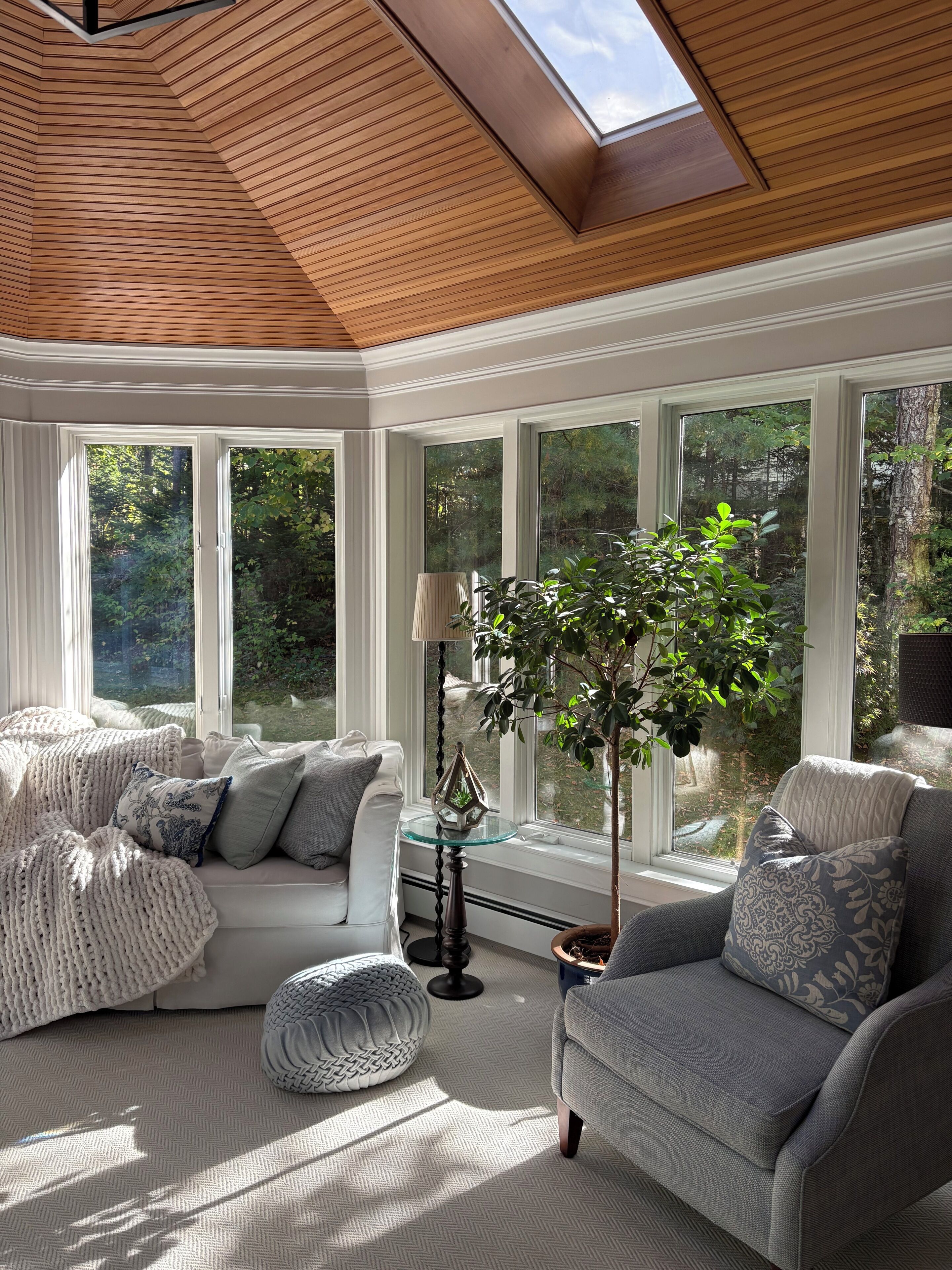 A sunlit room with a wooden ceiling, large windows, a white sofa, a grey armchair, and a potted plant.