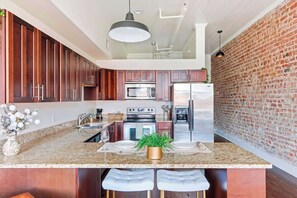 A modern kitchen with granite countertops, stainless steel appliances, and a brick accent wall. - Cozy Broughton Street Retreat | Walk to Everything (Savannah)