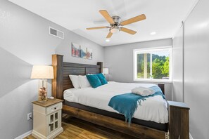 Ein Schlafzimmer mit Bett, Nachttisch, Deckenventilator, Fenster und einem an der Wand hängenden Kunstwerk.