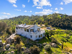 Una casa de dues plantes amb un balcó, envoltada d'un paisatge verd i unes muntanyes.