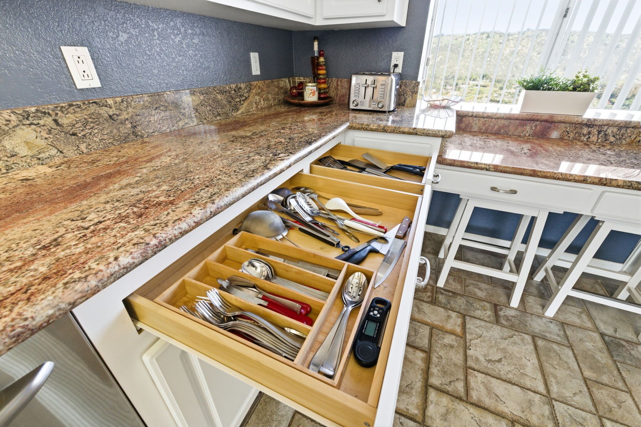 A kitchen with a granite countertop, a drawer organizer with utensils, a toaster, and a window with a view.