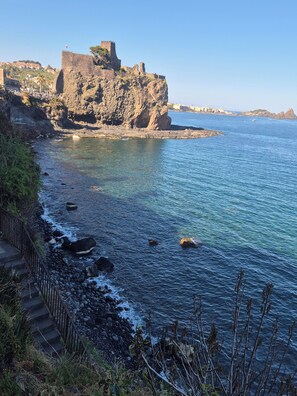 A coastal view with a rocky shoreline, a staircase leading down to the beach, and a historic fort on a cliff. - The A&G Choir   (Catania)