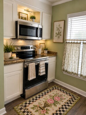 Cocina con armarios blancos, horno de acero inoxidable, microondas y ventana con cortinas florales.