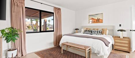 Une chambre moderne avec un grand lit, un banc, une plante en pot et une fenêtre avec des rideaux.