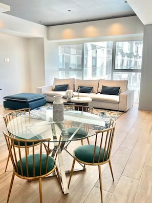 A modern living room with a glass dining table, green upholstered chairs, a white sofa with blue cushions, and large windows offering a city view. - Apartment in the Andares Financial District (Zapopan)