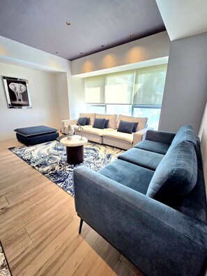 A modern living room with a grey sofa, a white sofa with blue cushions, a dark ottoman, and a patterned rug. - Apartment in the Andares Financial District (Zapopan)