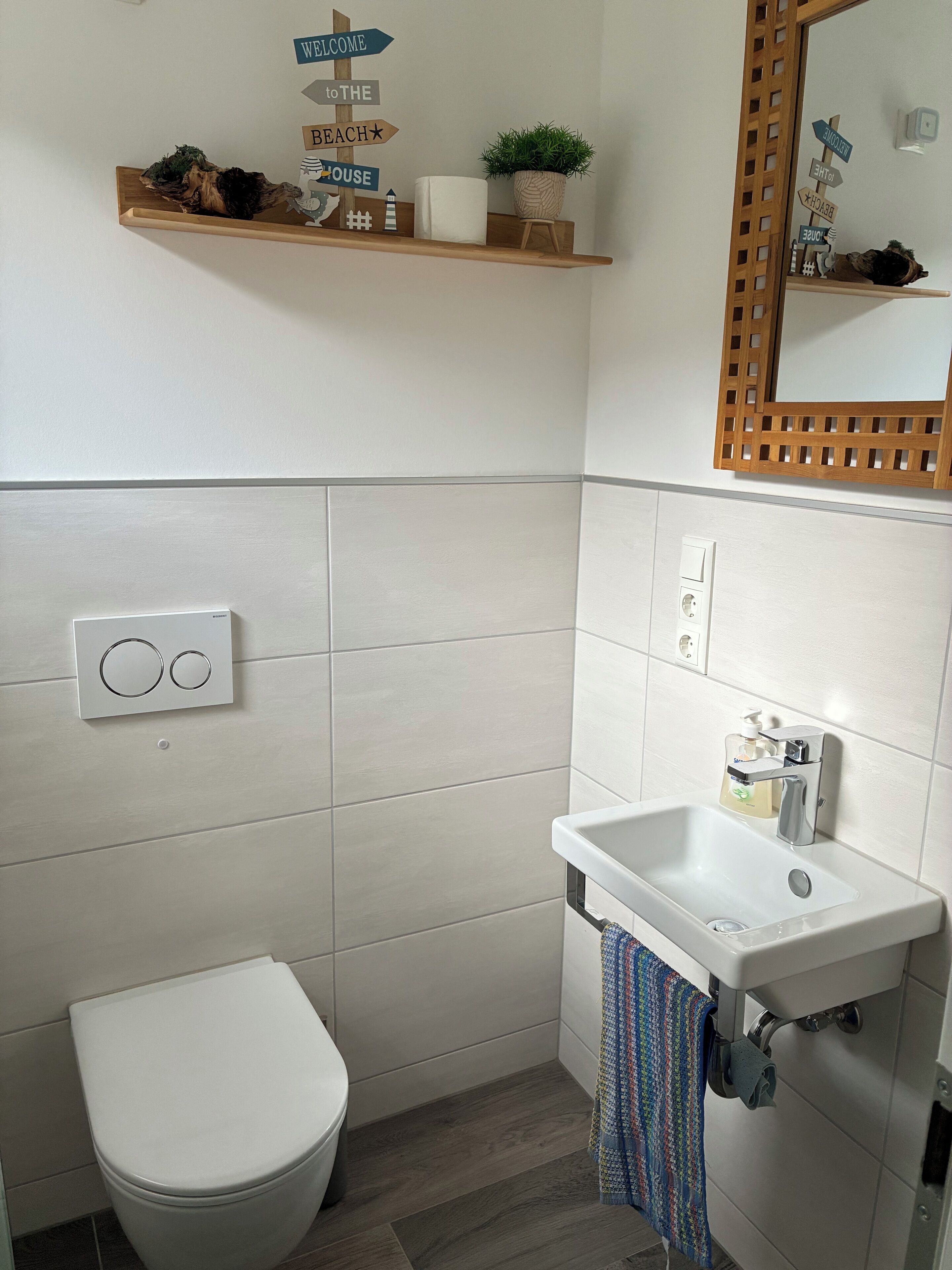 A bathroom with a toilet, a sink, and a mirror. There is a shelf with decorative items and a potted plant.