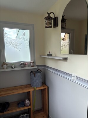 A room with a window displaying a floral pattern, a shelf with decorative items, a mirror, and a wooden bench with shoes. - Renovated farmhouse perfect for nature lovers and photo enthusiasts (Reurieth)
