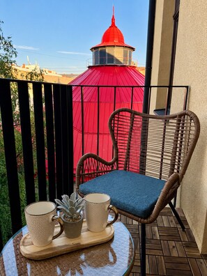 Ein Korbstuhl mit einem blauen Kissen auf einem Glastisch, eine Ablage mit zwei Tassen und einer kleinen Pflanze, sowie eine rote Struktur im Hintergrund.