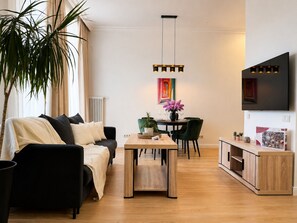 Ein modernes Wohnzimmer mit einer Couch, einem Esstisch und Stühlen. Ein Fernseher ist an der Wand montiert und in der Ecke steht eine Pflanze.