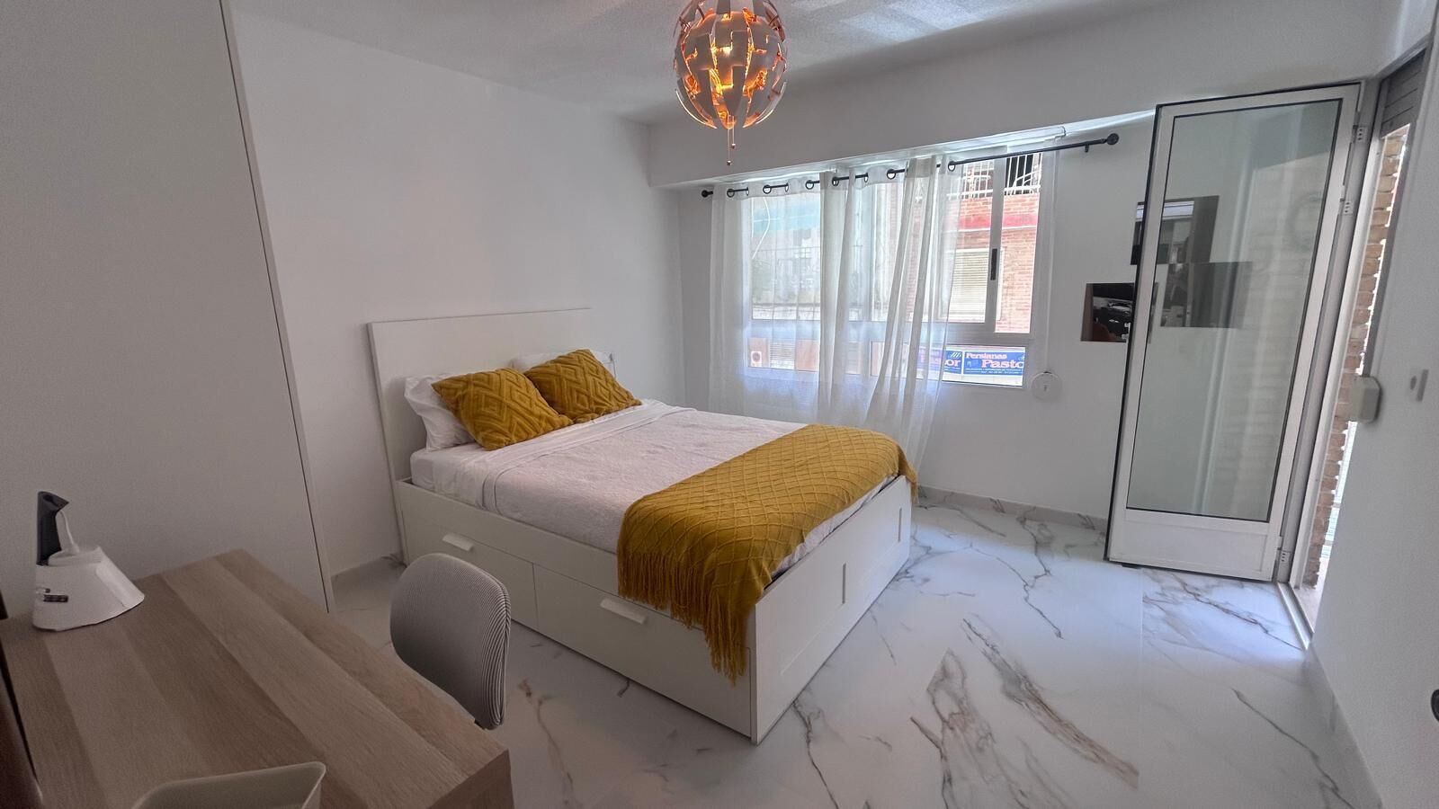 Une chambre à coucher comprenant un lit, un bureau, une chaise, une porte vitrée et une fenêtre avec des rideaux.