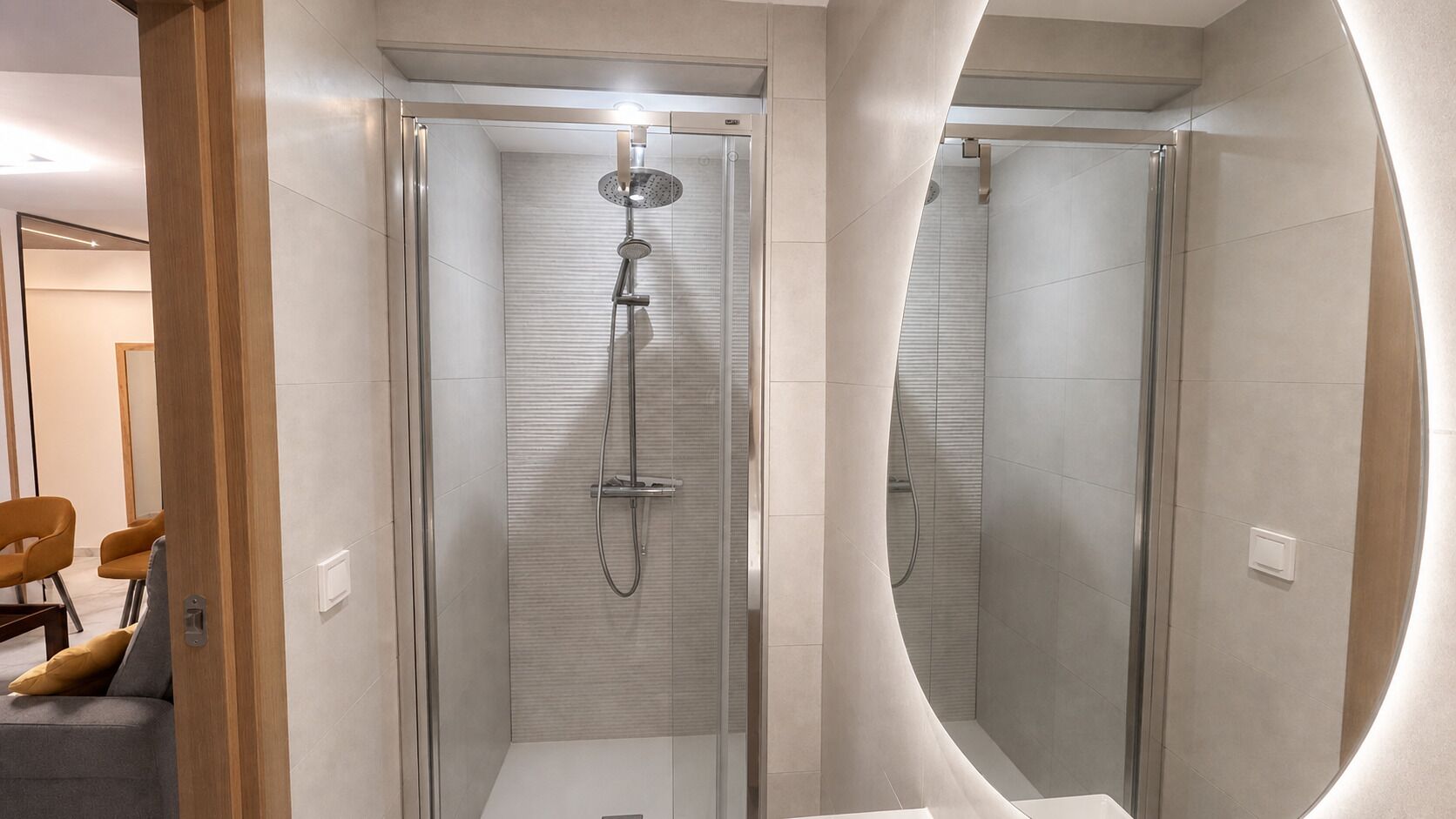 Et moderne badeværelse med et glasafskærmet brusebad, et spejl med træramme og et siddeområde med puder.