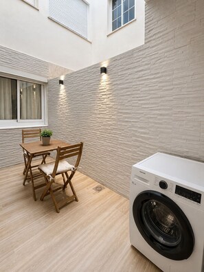 A compact laundry area with a washing machine, a wooden table with chairs, and a window with blinds. - Central Apartment | Within Walking Distance of the Train Station and Downtown (alicante)