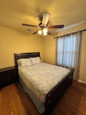 Ein Schlafzimmer mit einem Bett, einem Deckenventilator, einem Nachttisch und einem Fenster mit Vorhängen.