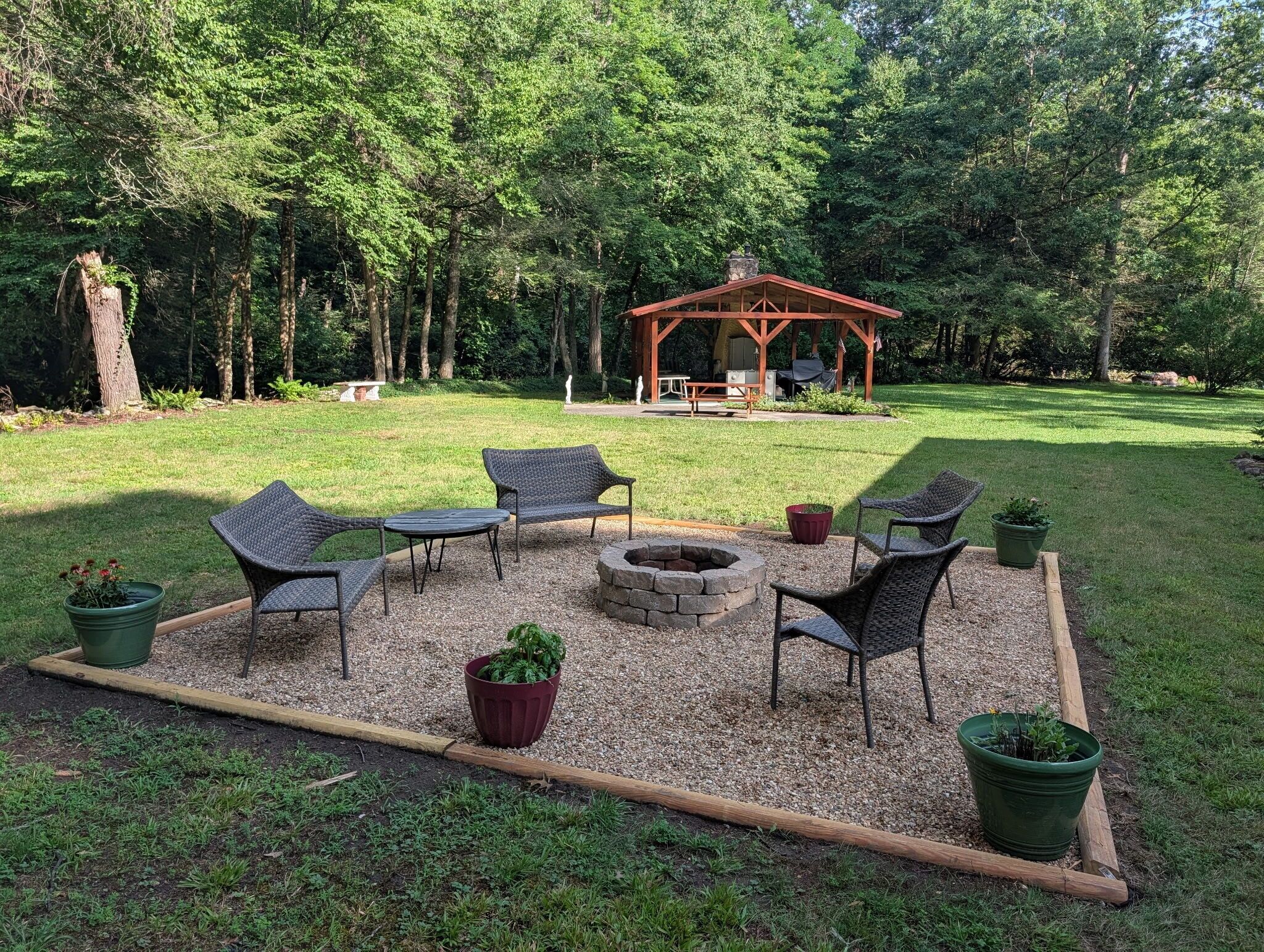 A gazebo with a fire pit, surrounded by wicker chairs and a table, set in a grassy area with trees in the background.