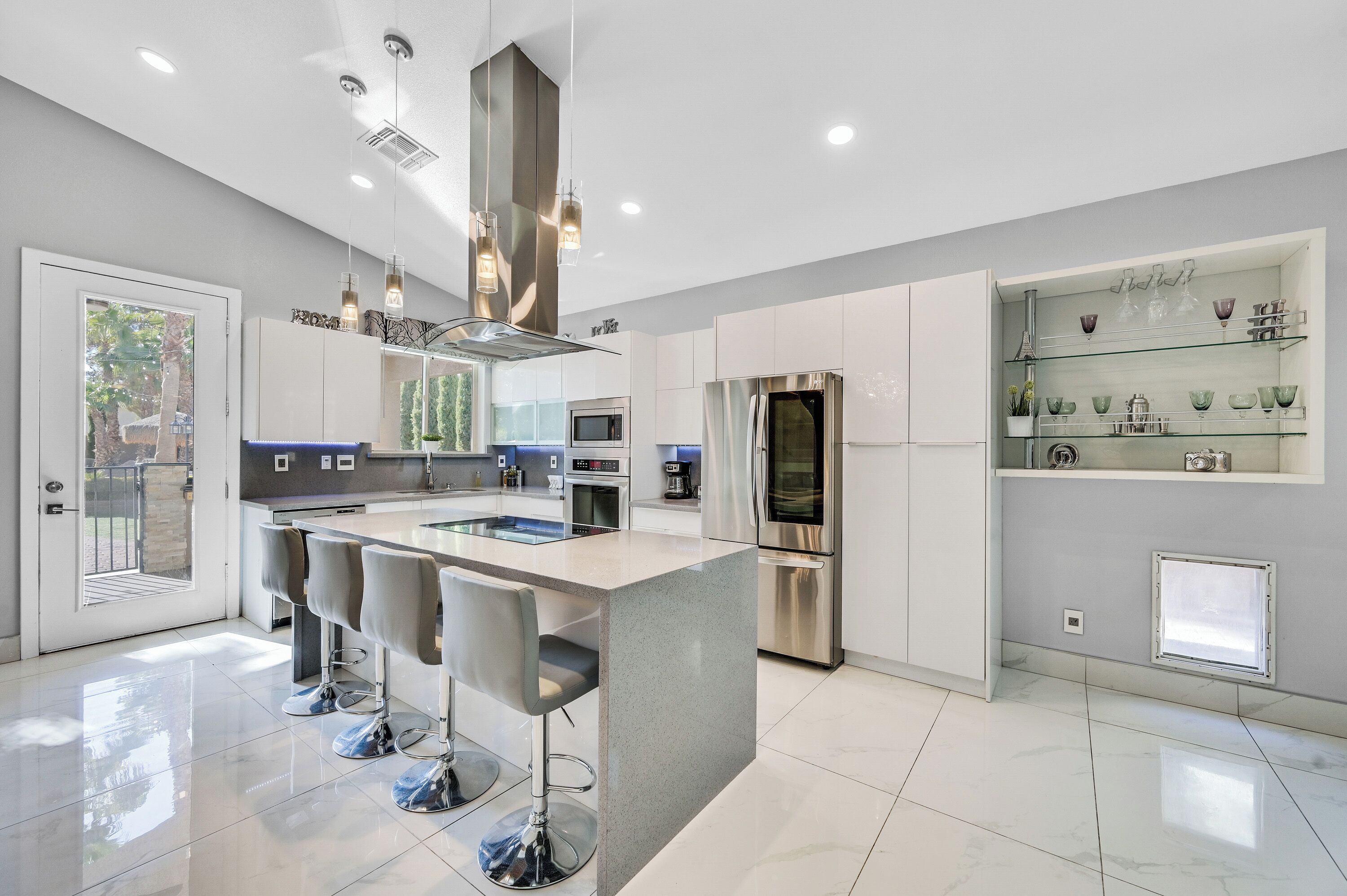 A modern kitchen with a central island, bar stools, stainless steel appliances, and a glass display shelf.