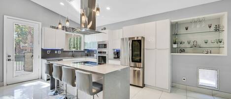 A modern kitchen with a central island, bar stools, stainless steel appliances, and a glass display shelf.