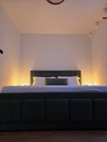Cama bem arrumada com cabeceira escura e roupa de cama branca, mesa de cabeceira com abajur e parede lisa com luminária circular no teto.