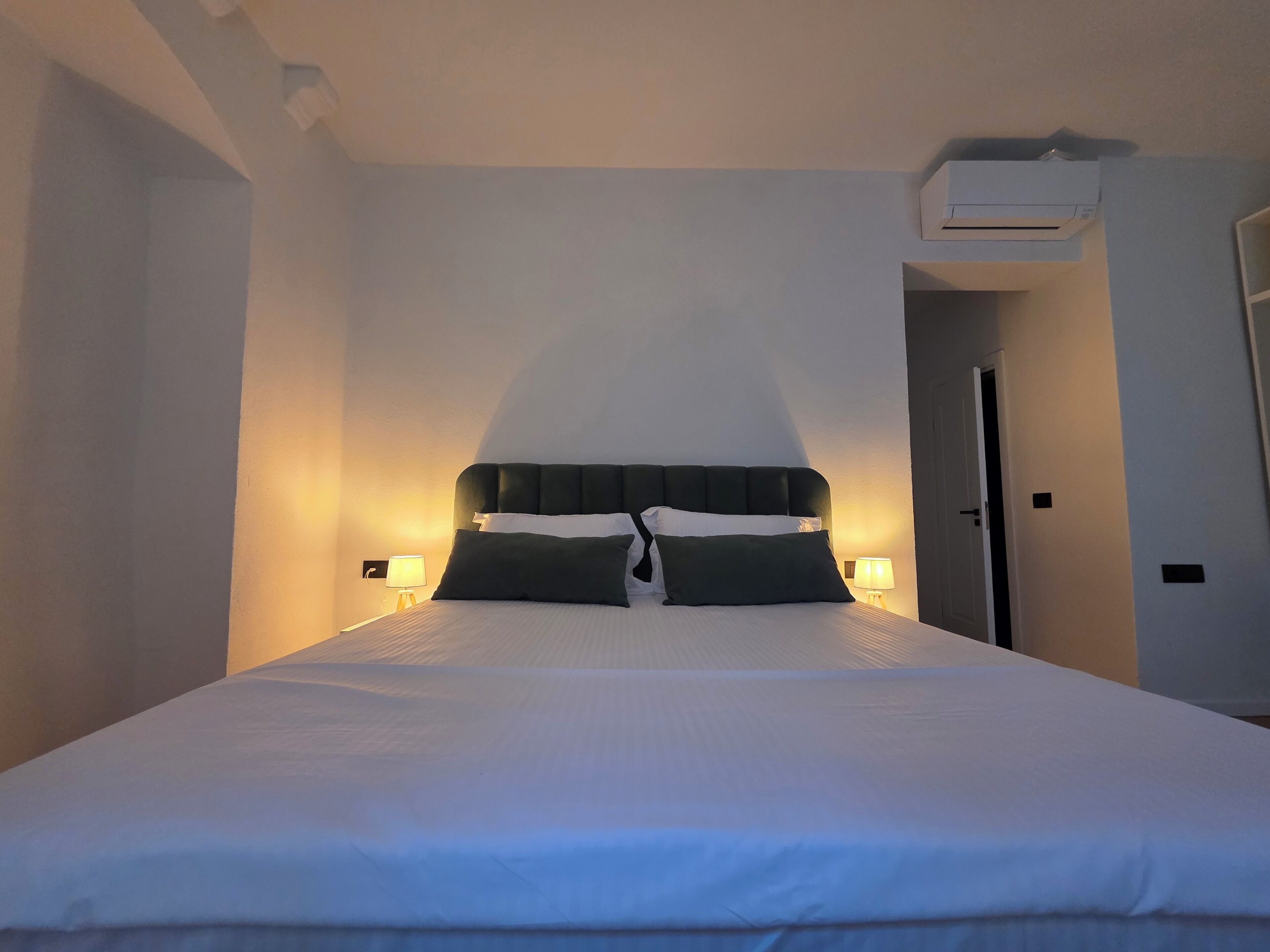 A hotel room with a large bed, two bedside lamps, a wall-mounted air conditioner, and a door.