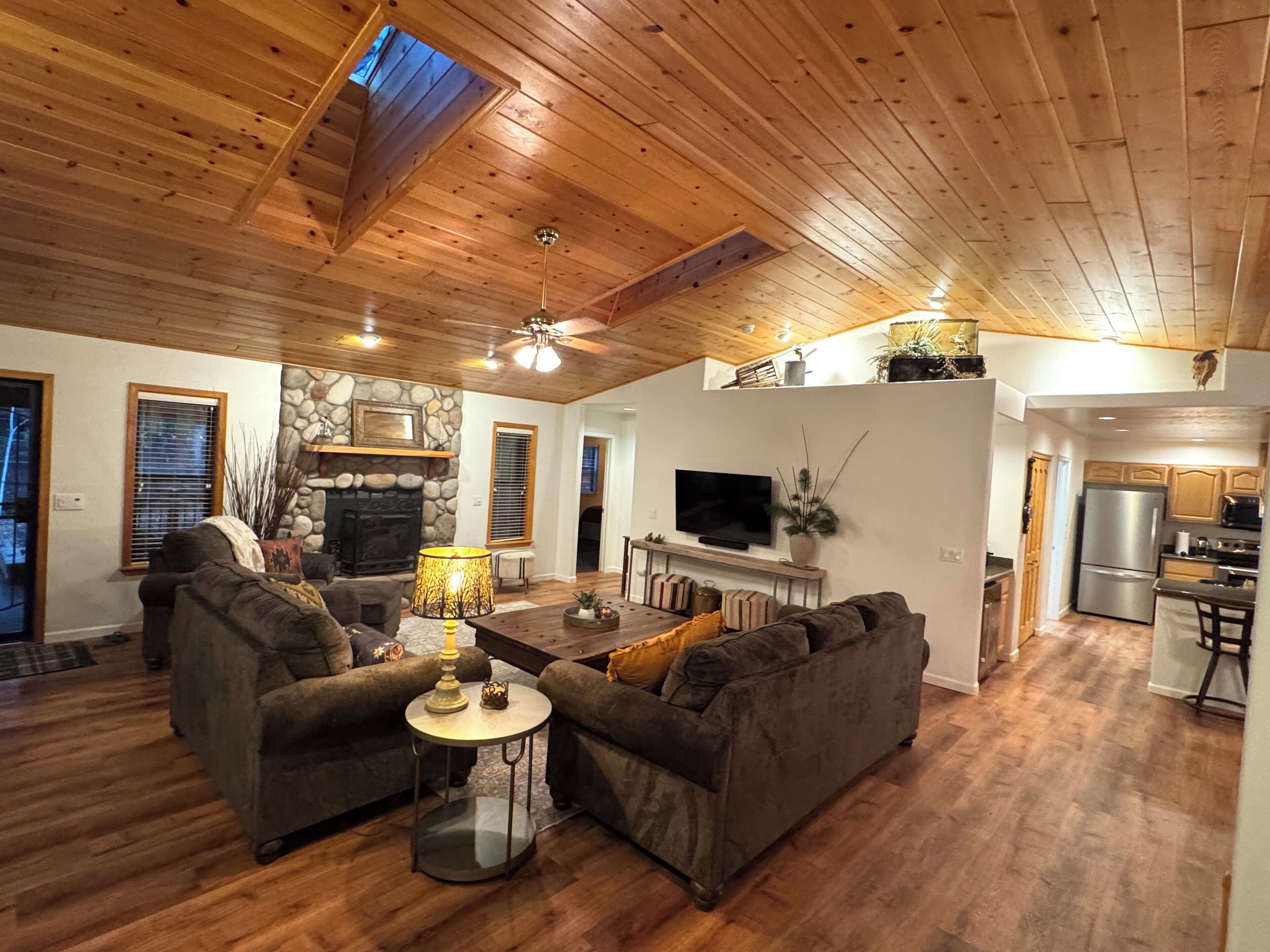 A spacious living room with a stone fireplace, wooden ceiling, and a kitchen area with modern appliances.