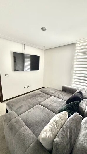 Ein Wohnzimmer mit einem großen grauen Sofa, einem flachbildfernseher an der Wand und einem Fenster mit Jalousien.