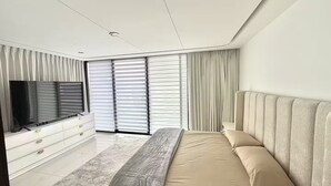 Ein modernes Schlafzimmer mit einem großen Bett, einem Fernseher und einem Fenster mit Jalousien.