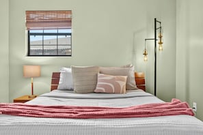 A bedroom with a bed, pillows, a bedside lamp, a window with blinds, and a wall-mounted light fixture. - Butler Beach Getaway | Tiki Bar + Fenced Yard! (St. Augustine)