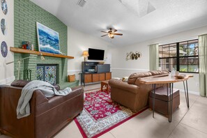 A living room with a fireplace, a television, a ceiling fan, and a dining area with a table and chairs. - Butler Beach Getaway | Tiki Bar + Fenced Yard! (St. Augustine)