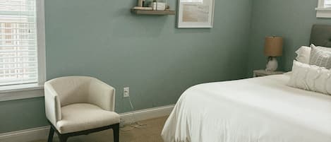 Um quarto com uma cama, uma cadeira, uma mesinha de cabeceira, uma luminária, uma estante com objetos decorativos e um quadro na parede.