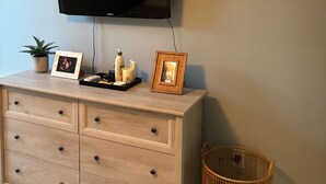 A room with a flat-screen TV mounted on the wall, a wooden dresser with drawers, a small potted plant, a framed picture, and a wicker basket. - Casa Sul Lago Spacious 5-bedroom house in charming Delavan w/AC Firepit Deck (Delavan)
