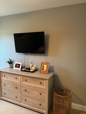 Ein Zimmer mit flachbildfernseher an der Wand, einer Holzkommode mit Schubladen, einer kleinen Topfpflanze, einem gerahmten Bild und einem Korbwägelchen.