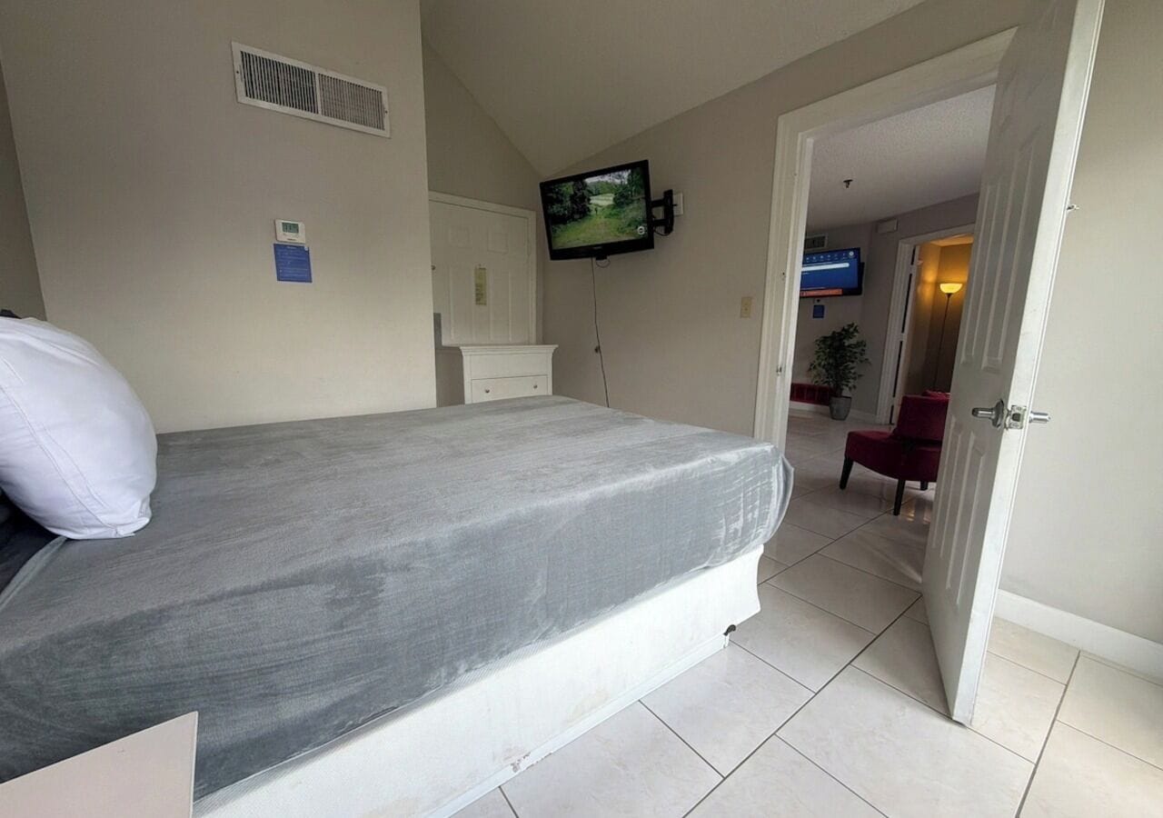 A bedroom with a bed, a television mounted on the wall, a white dresser, and a door leading to another room.
