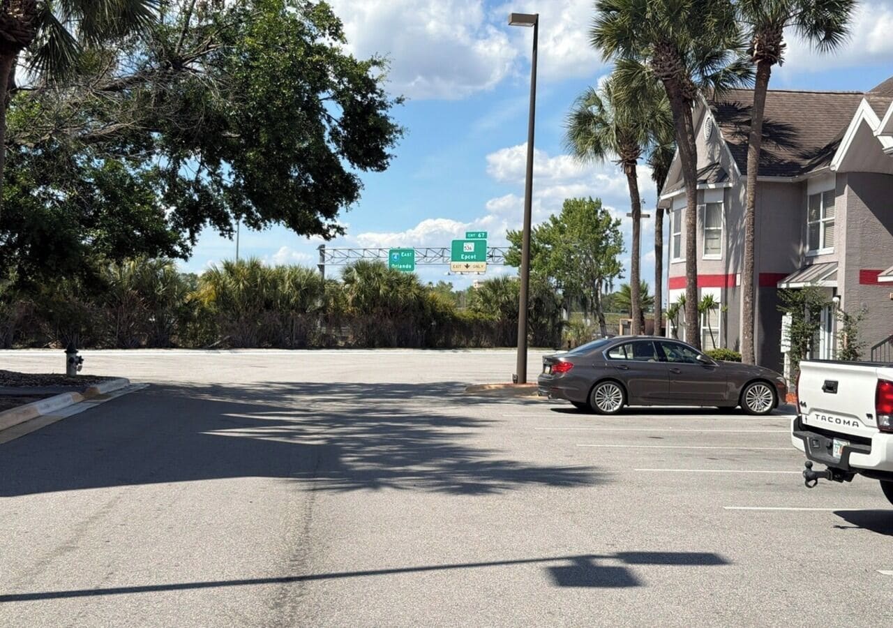 Un estacionamiento con un coche, un camión, árboles y un edificio con un cartel que indica indicaciones para Florida Este y la salida 7.