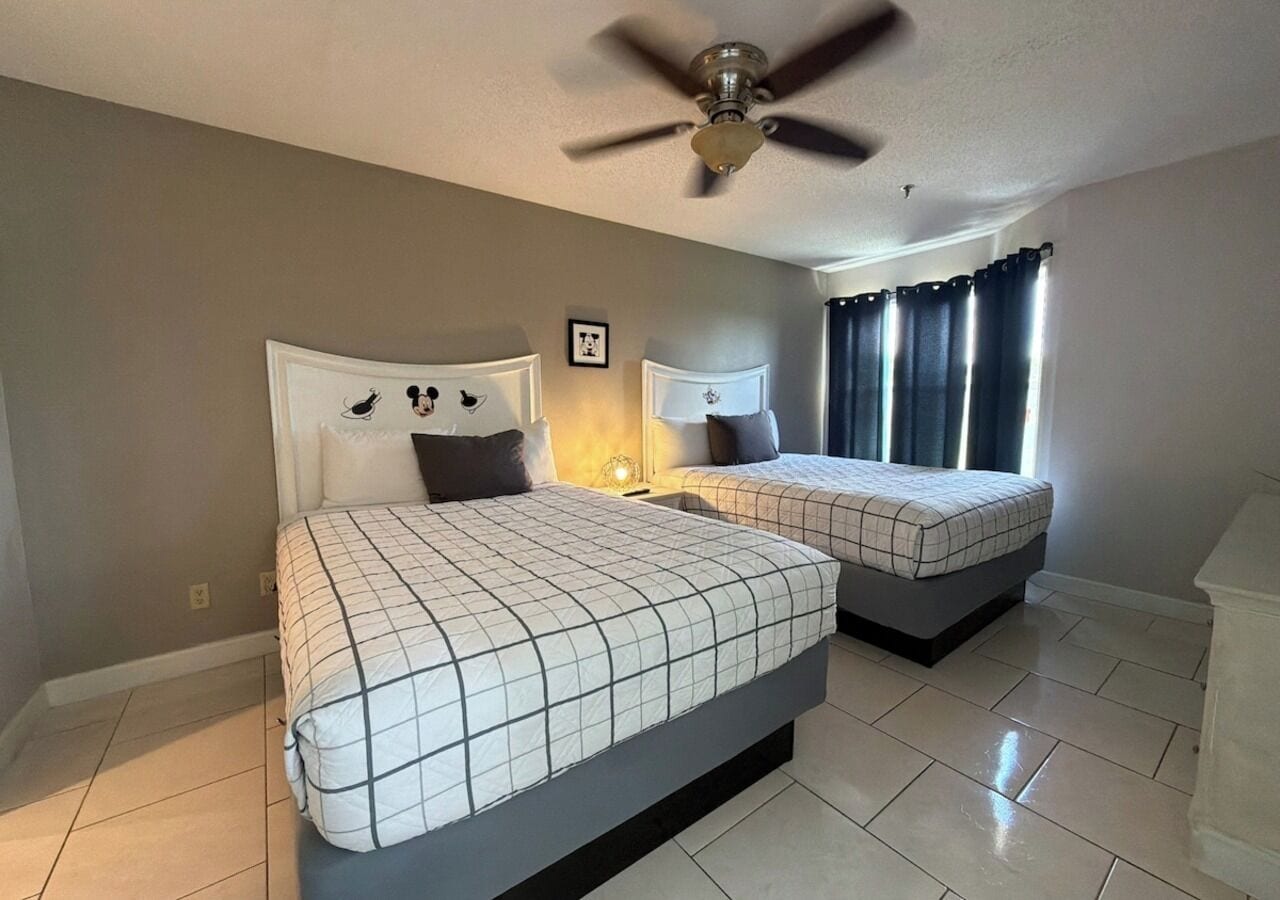 Un dormitorio con dos camas, un ventilador de techo, una ventana con cortinas y un cuadro en la pared.