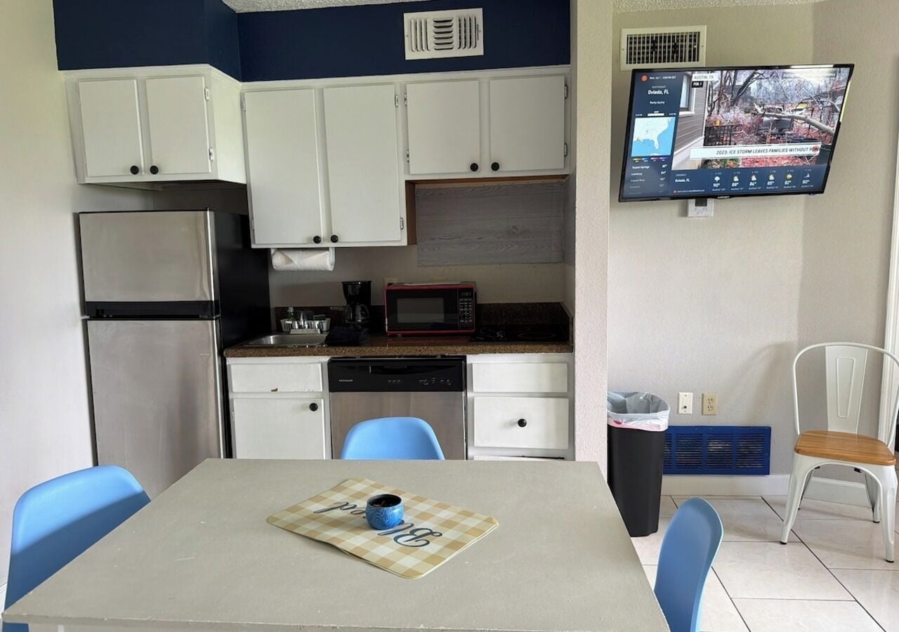 A compact kitchen with white cabinets, a microwave, and a refrigerator. A television is mounted on the wall displaying a weather forecast.