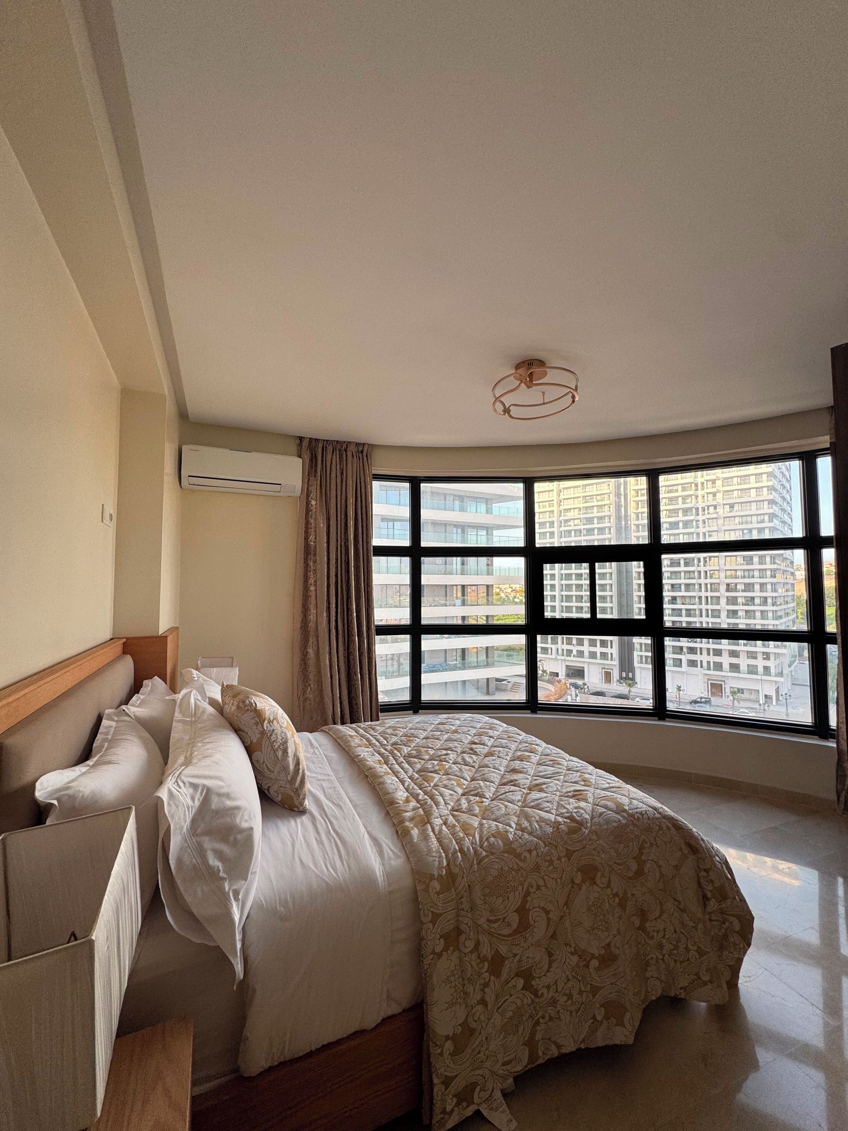 A bedroom with a large window, a bed with a patterned bedspread, a bedside table, and an air conditioning unit.