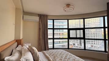 Een slaapkamer met een groot raam, een bed met een bedspreide met dessin, een nachtkastje en een airconditioning.