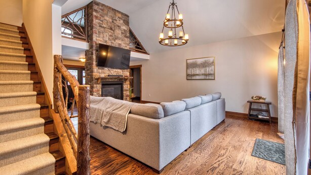 A modern living room with a stone fireplace, a grey sofa, wooden flooring, and a staircase with a wooden handrail. - Jay Brook Lodge | Sleeps 16 | Hot Tub | Game Room (Jay)