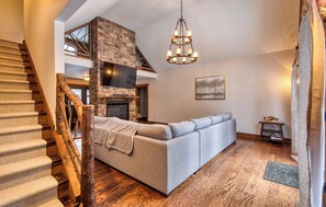 Un salon moderne avec une cheminée en pierre, un canapé gris, un plancher en bois et un escalier doté d’une rampe en bois.