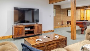 A living room with a TV, wooden cabinet, pool table, and a coffee table with game controllers. - Jay Brook Lodge | Sleeps 16 | Hot Tub | Game Room (Jay)