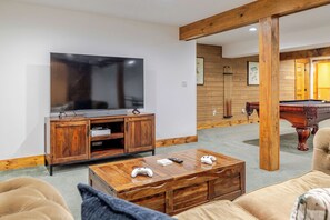 A living room with a TV, wooden cabinet, pool table, and a coffee table with game controllers. - Jay Brook Lodge | Sleeps 16 | Hot Tub | Game Room (Jay)
