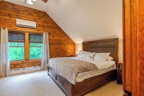 Une chambre avec un plafond en pente, des murs en bois, un lit avec une tête de lit, une fenêtre avec des stores et un appareil de climatisation.