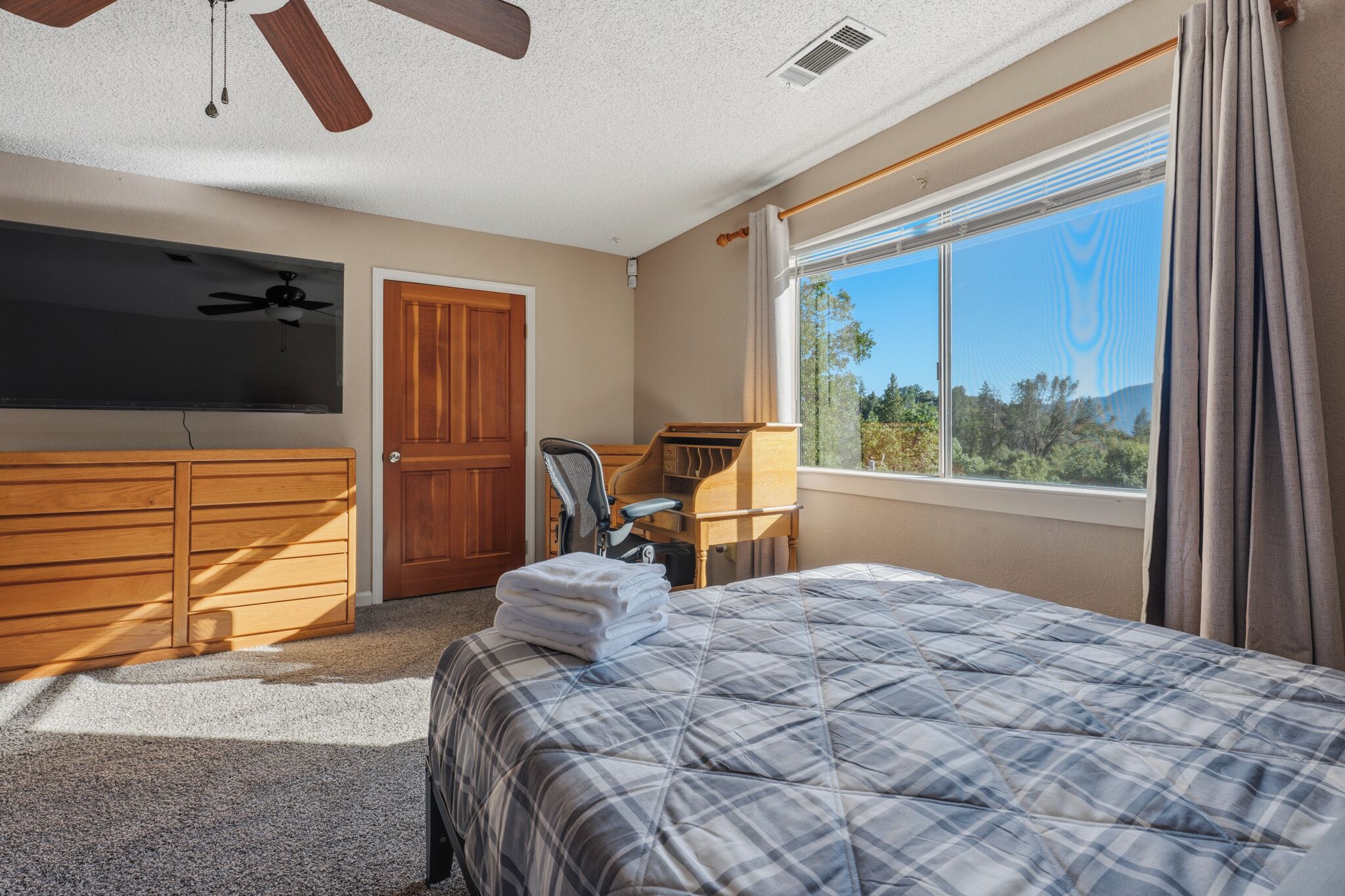 Une chambre à coucher comprenant un lit, un bureau, une chaise, un téléviseur, un ventilateur de plafond et une fenêtre avec des rideaux.