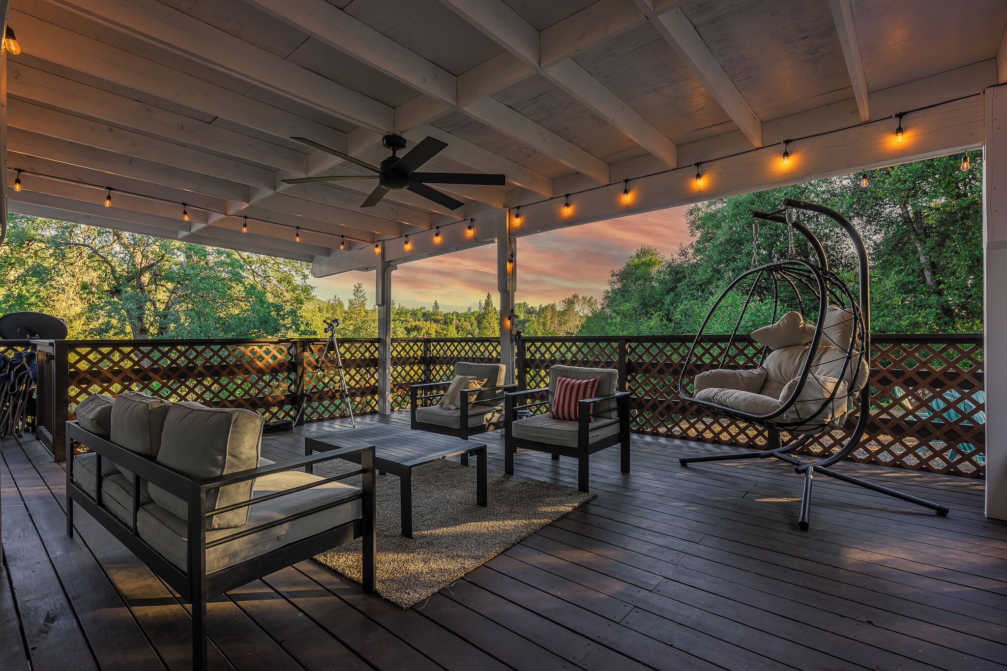 A covered outdoor deck with wooden flooring, a ceiling fan, string lights, a hanging chair, and a wooden lattice fence.