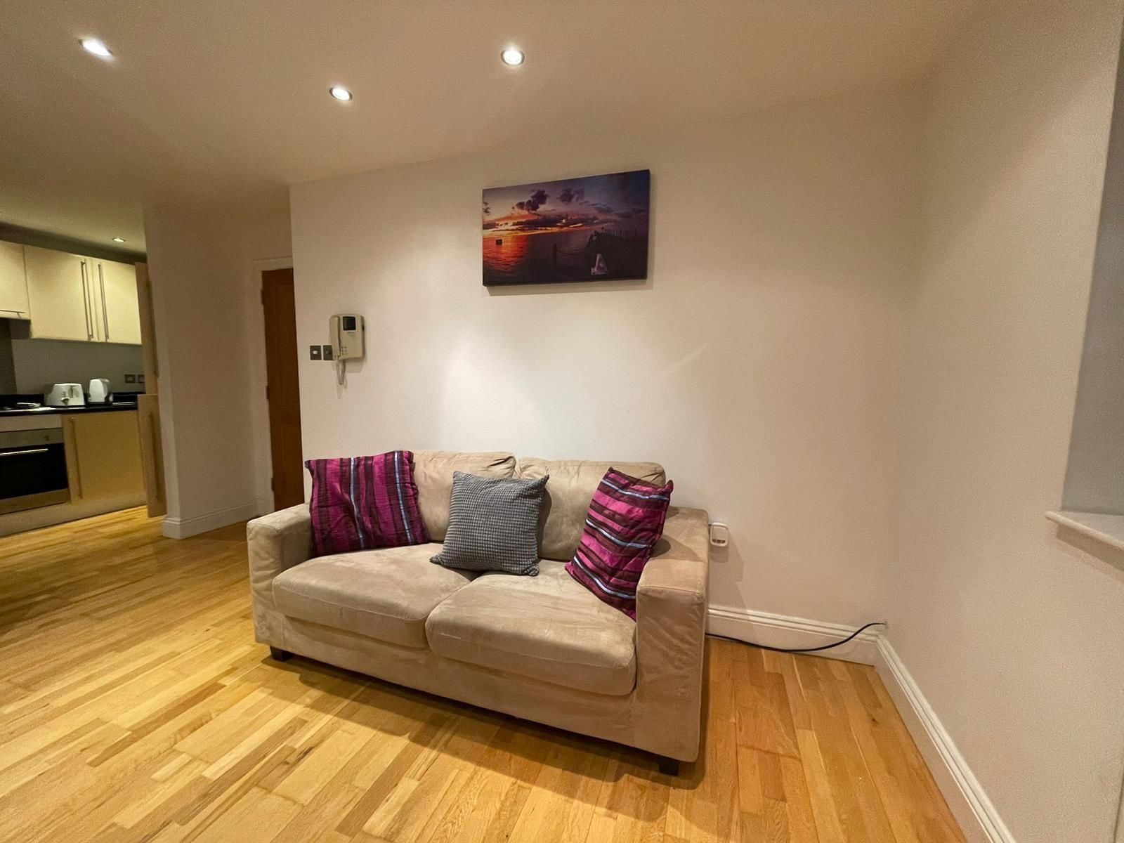 A living room with a beige sofa, two pillows, a painting on the wall, and a kitchen in the background.